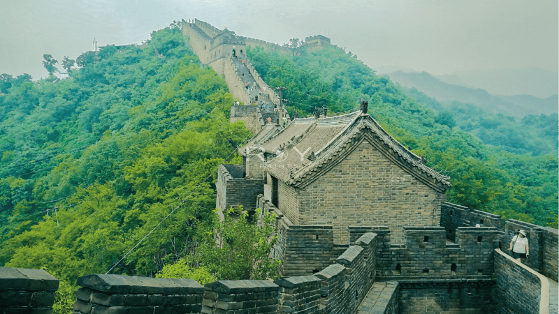 Stone ramparts of the Great Wall wind over green hills with tourists climbing the iconic landmark.