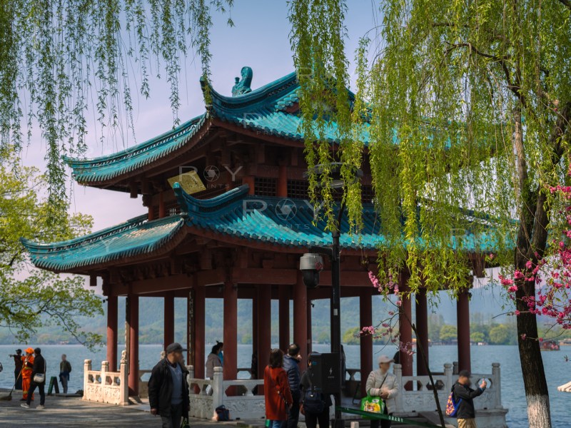 Traditional Chinese pavilion with a blue roof at West Lake, surrounded by willow trees, flowers, and visitors.